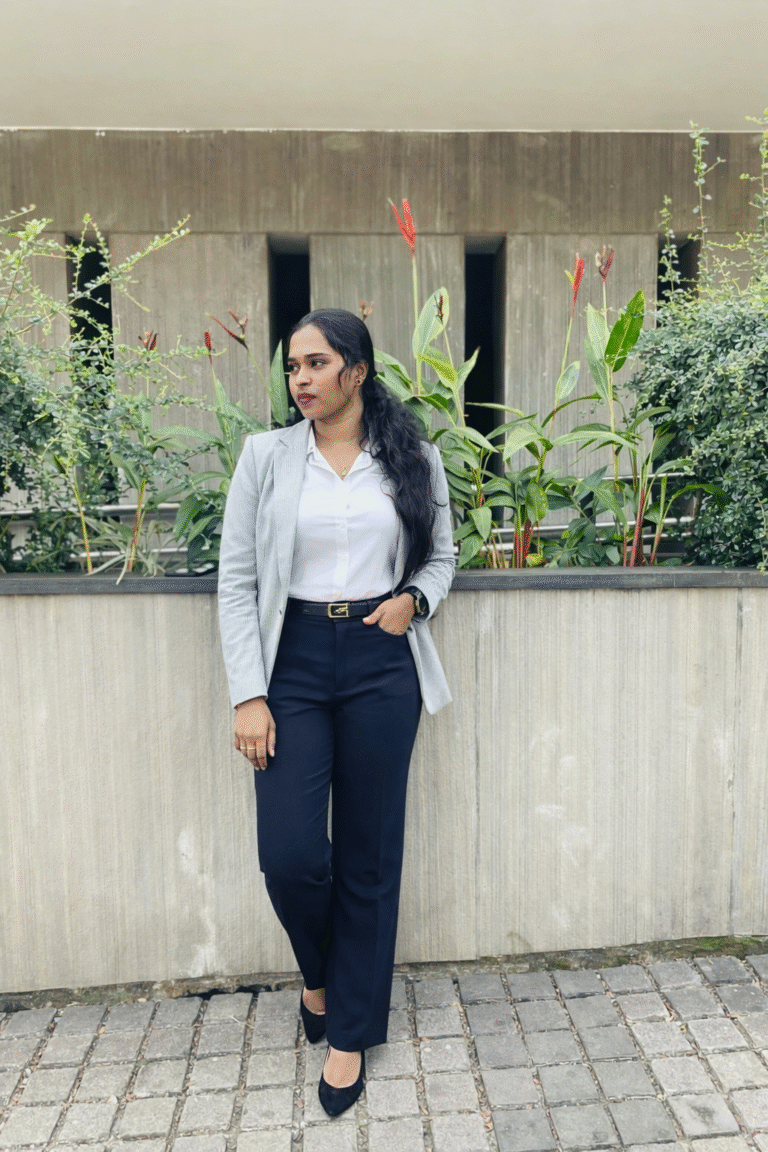 Aswathi.sk standing confidently in gray blazer,white shirt,and navy trousers against a modern architectural background with greenery.